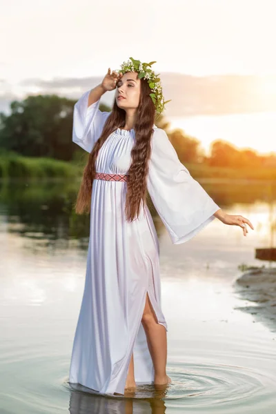 Gyönyörű Fekete hajú lány fehér vintage ruhát és a tó vize állandó virágok koszorút. Napsütés fáklyát. — Stock Fotó