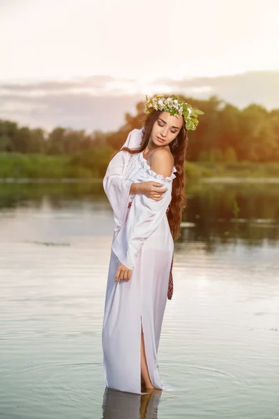 Beautiful black haired girl in white vintage dress and wreath of flowers standing in water of lake. Sun flare. — Stock Photo, Image