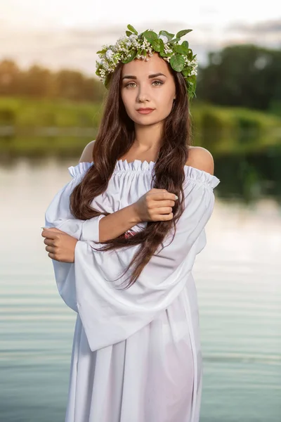 Beautiful black haired girl in white vintage dress and wreath of flowers standing in water of lake. Sun flare.
