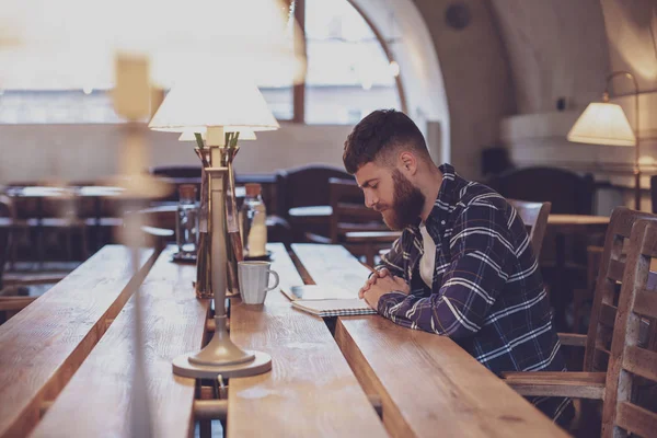 Young bearded businessman sits in cafe, home at table and writes
