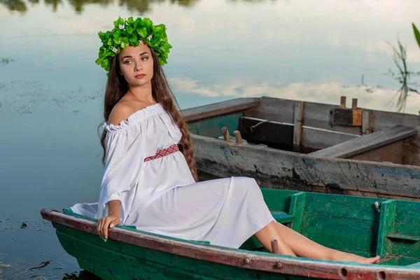 Junge Frau mit Blumenkranz auf dem Kopf, die es sich bei Sonnenuntergang auf einem Boot auf dem Fluss gemütlich macht. Konzept der weiblichen Schönheit, Ruhe im Dorf — Stockfoto