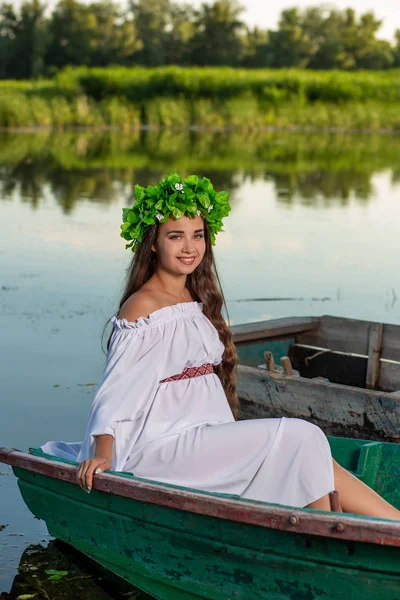 Fiatal nő Virág Koszorú a fején, pihentető a hajón folyó naplementekor. Koncepció a női szépség, a többi a faluban — Stock Fotó