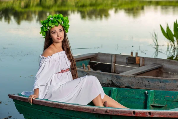 Fiatal nő Virág Koszorú a fején, pihentető a hajón folyó naplementekor. Koncepció a női szépség, a többi a faluban — Stock Fotó