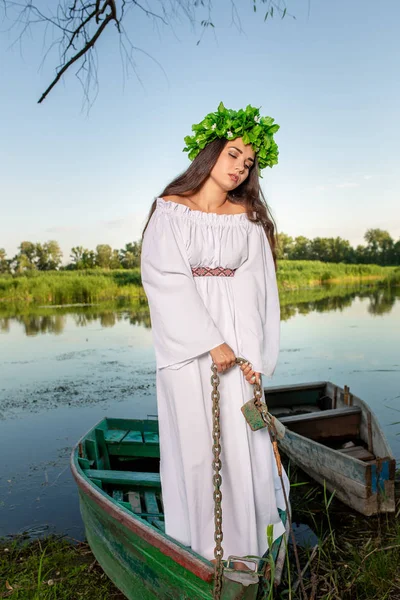 Fiatal nő Virág Koszorú a fején, pihentető a hajón folyó naplementekor. Koncepció a női szépség, a többi a faluban — Stock Fotó