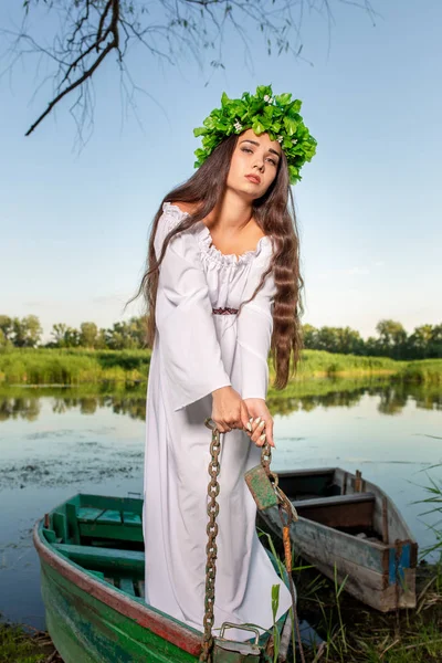 Young woman with flower wreath on her head, relaxing on boat on river at sunset. Concept of female beauty, rest in the village