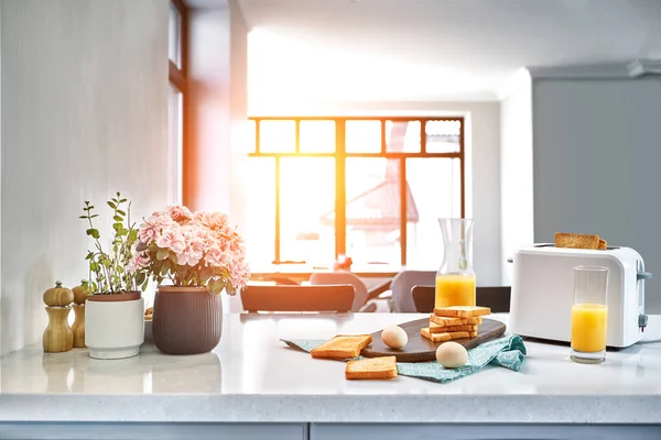 Тостер со свежими тостами, яйцами и стаканом апельсинового сока на светлом кухонном столе. Солнечная вспышка — стоковое фото