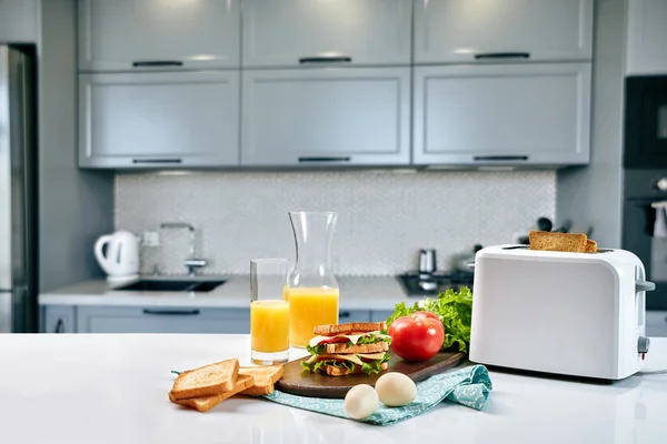 Weißer elektrischer Toaster mit Orangensaft und Sandwich auf dem Küchentisch. Frühstück in der gemütlichen Küche. — Stockfoto