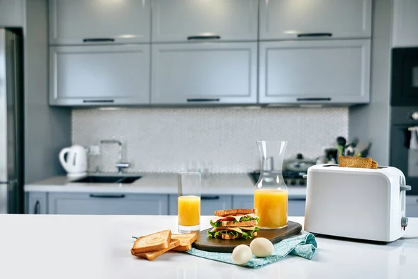 Weißer elektrischer Toaster mit Orangensaft und Sandwich auf dem Küchentisch. Frühstück in der gemütlichen Küche. — Stockfoto