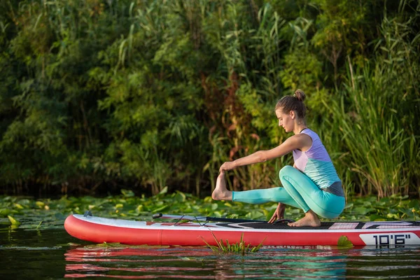 Giovane donna stanno facendo yoga su uno stand up paddle board SUP su un bellissimo lago o fiume — Foto Stock