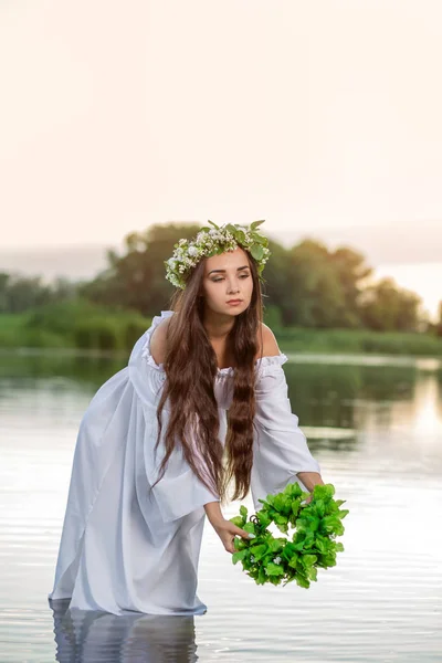 Beautiful woman on the river bank. Rustic woman with a wreath in a transparent dress