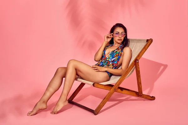 Sommer-Lifestyle-Mode Porträt einer jungen atemberaubenden Frau auf einem Liegestuhl, rosa Hintergrund, Studioaufnahme. mit stylischer Sonnenbrille, Bikini. — Stockfoto