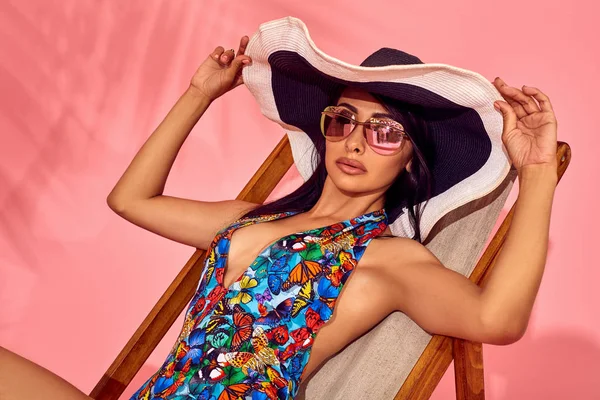 Zomer levensstijl mode portret van jonge prachtige vrouw op een lounge stoel, roze achtergrond, studio opname. Dragen van een stijlvolle zonnebril, bikini. — Stockfoto