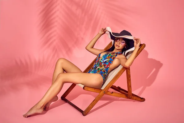 Summer lifestyle fashion portrait of young stunning woman on a lounge chair, pink background, studio shot. Wearing stylish sunglasses, bikini. — Stock Photo, Image