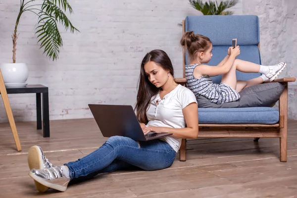 Mãe trabalhando com laptop e linda menina deitada na poltrona e jogando jogos no smartphone — Fotografia de Stock
