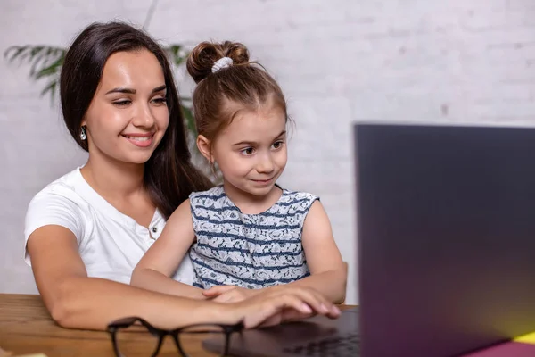 Привлекательная молодая женщина и ее маленькая милая дочь сидят за столом и веселятся, выполняя домашнее задание вместе . — стоковое фото