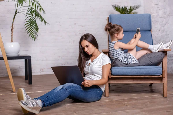 Красивая брюнетка мама работает с ноутбуком и симпатичная маленькая девочка лежит на кресле и играет в игры на смартфоне — стоковое фото