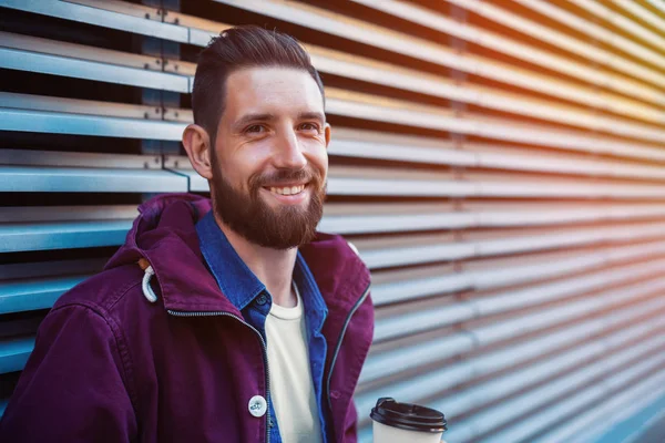 Όμορφος νεαρός άνδρας στο μωβ χειμερινό μπουφάν πίνοντας καφέ. Ο άνθρωπος που πίνει καφέ στο δρόμο. Ήλιος φωτοβολίδα — Φωτογραφία Αρχείου