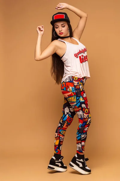 Una giovane donna carina vestibilità attraente indossa leggings multicolori, jersey bianco e cappuccio. Immagine a figura intera, sfondo giallo studio — Foto Stock