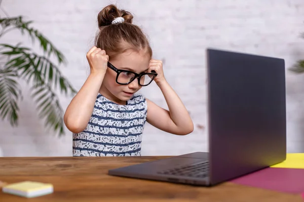 Милая маленькая девочка сидит за столом со своим черным ноутбуком и носит очки дома — стоковое фото
