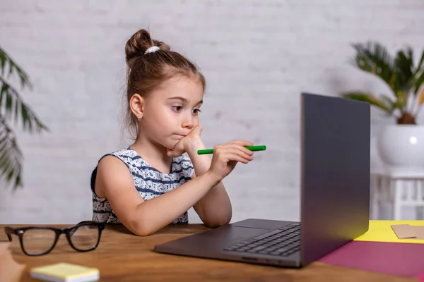 Милая маленькая девочка сидит за столом со своим ноутбуком и блокнотом, в очках — стоковое фото