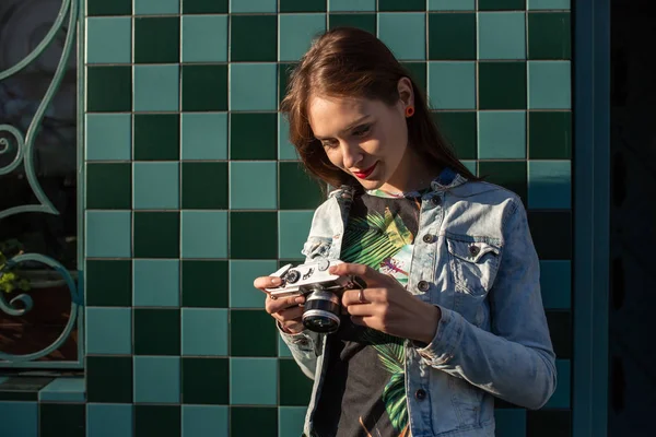 Modelo engraçado legal da menina com câmera de filme retro vestindo uma jaqueta de ganga, cabelo escuro ao ar livre sobre a parede da cidade em um fundo de gaiola — Fotografia de Stock