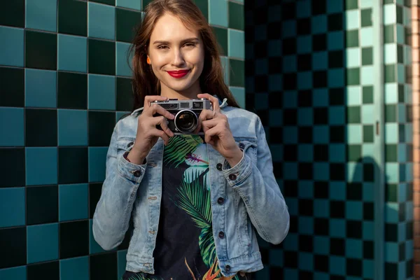 Крутая смешная девушка модель с ретро-кинокамерой в джинсовой куртке, темные волосы на открытом воздухе над городской стеной на фоне клетки — стоковое фото