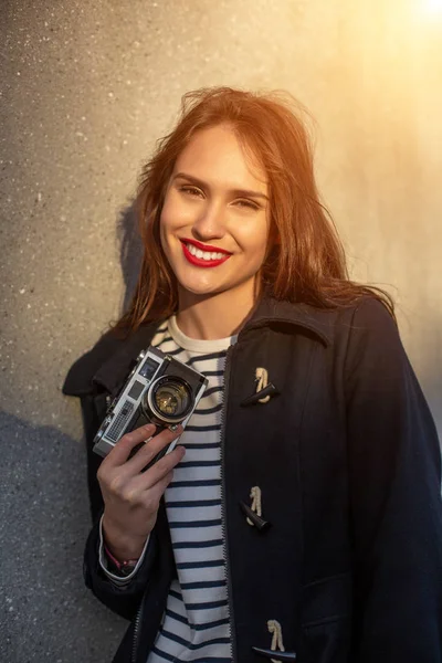 Smiling female photographer in jacket standing in front of wall ready to make new photo. Sun flare