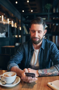 Genç işadamı, kafe ve kullanım smartphone masada oturan bir kot gömlek giymiş sakallı. Adam Gadget'ı kullanma.