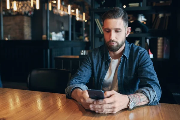 Joven hombre de negocios barbudo, vestido con una camisa de mezclilla, sentado en la mesa en la cafetería y el uso de teléfono inteligente. Hombre usando gadget . — Foto de Stock