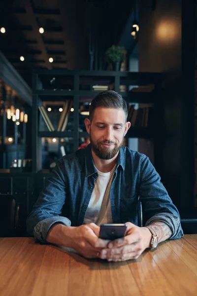 Joven hombre de negocios barbudo, vestido con una camisa de mezclilla, sentado en la mesa en la cafetería y el uso de teléfono inteligente. Hombre usando gadget . — Foto de Stock
