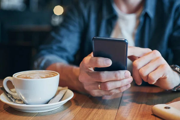Las manos del hombre en la camisa de mezclilla se deslizan con el dedo en la pantalla su teléfono inteligente, cerca de la taza con café en la mesa de madera — Foto de Stock