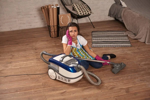 Una joven ama de casa sentada apoyada en una aspiradora cansada de las tareas domésticas . — Foto de Stock