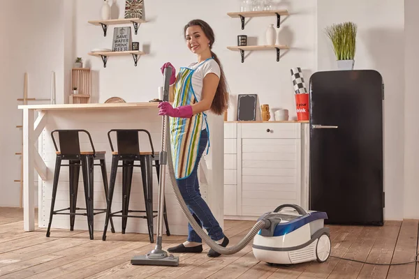 Giovane donna che utilizza l'aspirapolvere nel pavimento della cucina di casa, facendo compiti di pulizia e faccende, interni meticolosi . — Foto Stock