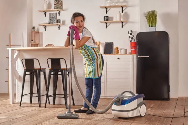 Young woman using vacuum cleaner in home kitchen floor, doing cleaning duties and chores, meticulous interior.