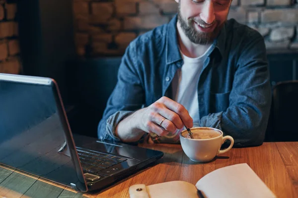 Hombre joven bebiendo café en la cafetería de la ciudad durante el almuerzo y trabajando en el ordenador portátil — Foto de Stock