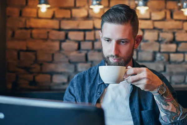 Hombre joven bebiendo café en la cafetería de la ciudad durante el almuerzo y trabajando en el ordenador portátil — Foto de Stock