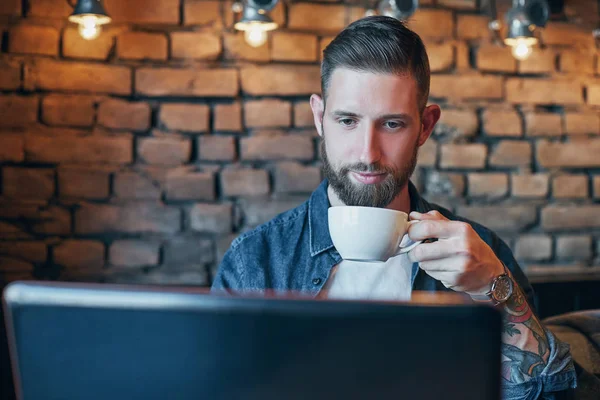 Hombre joven bebiendo café en la cafetería de la ciudad durante el almuerzo y trabajando en el ordenador portátil — Foto de Stock