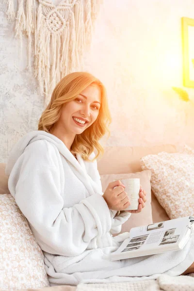 Young woman in white terry robe is drinking coffee and reading magazine or book in bedroom.