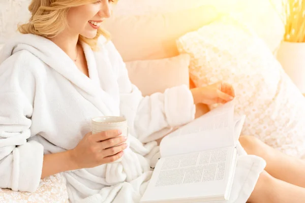 Young woman in white terry robe is drinking coffee and reading magazine or book in bedroom.