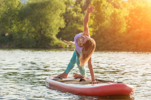 Young woman doing yoga on sup board with paddle. Yoga pose, side view - concept of harmony with the nature.