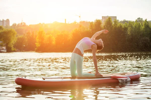 Young woman doing yoga on sup board with paddle. Yoga pose, side view - concept of harmony with the nature.