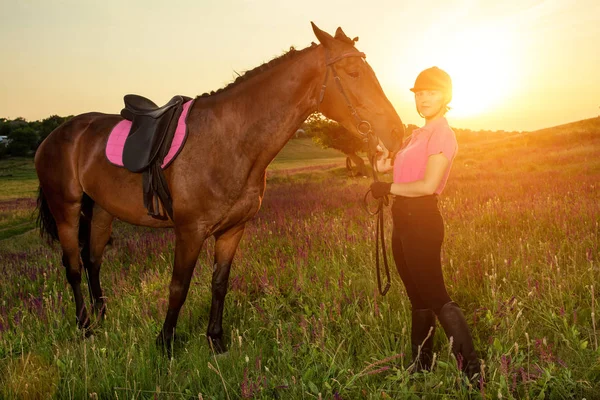 Beautiful smiling girl jockey stand next to her brown horse wearing special uniform on a sky and green field background on a sunset. — Stock Photo, Image