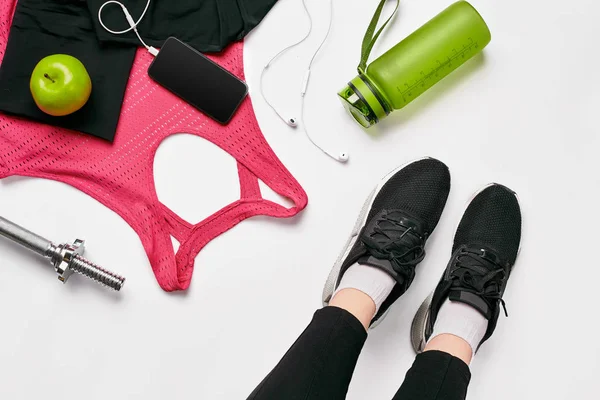 Mädchenbeine in schwarzen Turnschuhen sitzen auf einer Matte, umgeben von Handy, Kopfhörern, Handtuch und einem Shaker im Fitnessclub. Ansicht von oben. — Stockfoto