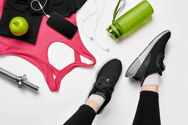 Nogi dziewczyny na sobie czarne Adidasy siedzi na matę, otoczony przez telefon komórkowy, słuchawki, ręcznik i shakerze w klubie fitness. Widok z góry. — Zdjęcie stockowe