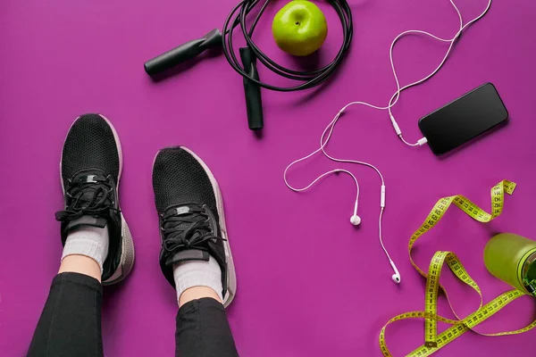 Jambes de filles portant des baskets noires assises sur un tapis entouré d'un téléphone portable, écouteurs, serviette et un shaker dans le club de fitness. Vue du dessus . — Photo