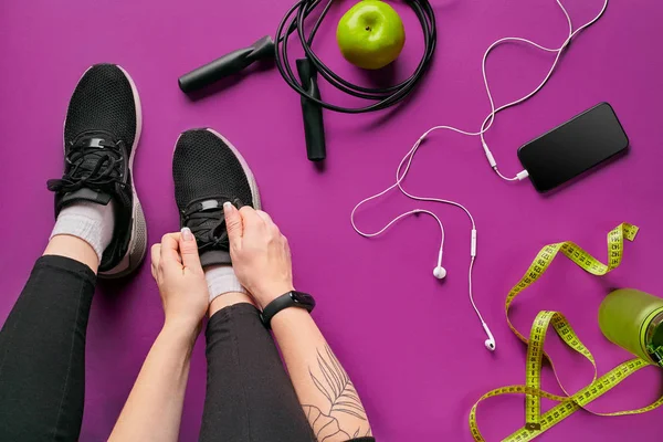 Junge Frau schnürt Turnschuhe und bereitet sich auf das Training vor. Wasserflasche, Yogamatte, Telefon, Kopfhörer auf lila Hintergrund flache Lageoberseite. — Stockfoto