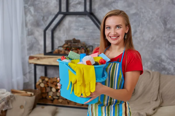 Uma mulher de limpeza está de pé na sala de estar segurando um balde azul preenchido com produtos químicos e instalações para arrumar em sua mão — Fotografia de Stock