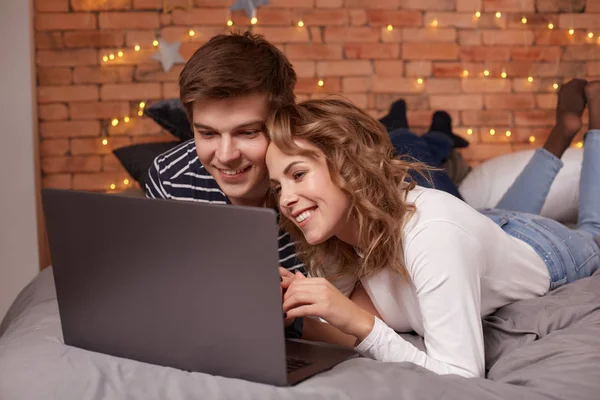 Sonriendo joven pareja hombre y mujer acostados en la cama y viendo algo en el portátil — Foto de Stock