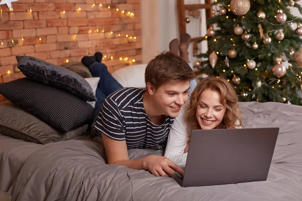 Sonriendo joven pareja hombre y mujer acostados en la cama y viendo algo en el portátil — Foto de Stock