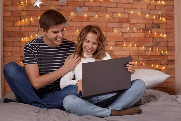 Sonriendo joven pareja hombre y mujer sentado en la cama y utilizando su ordenador portátil — Foto de Stock